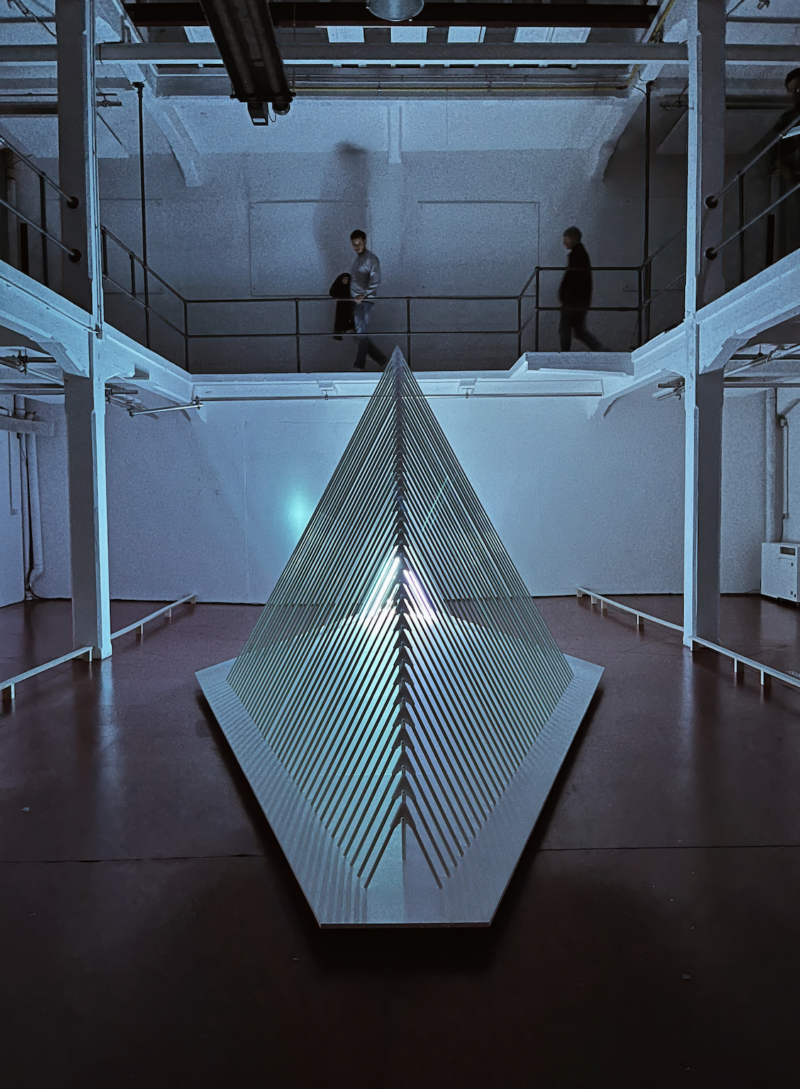 Photography of a kinectic installation in a shape of a pyramid in a two floors room. Blue lights.