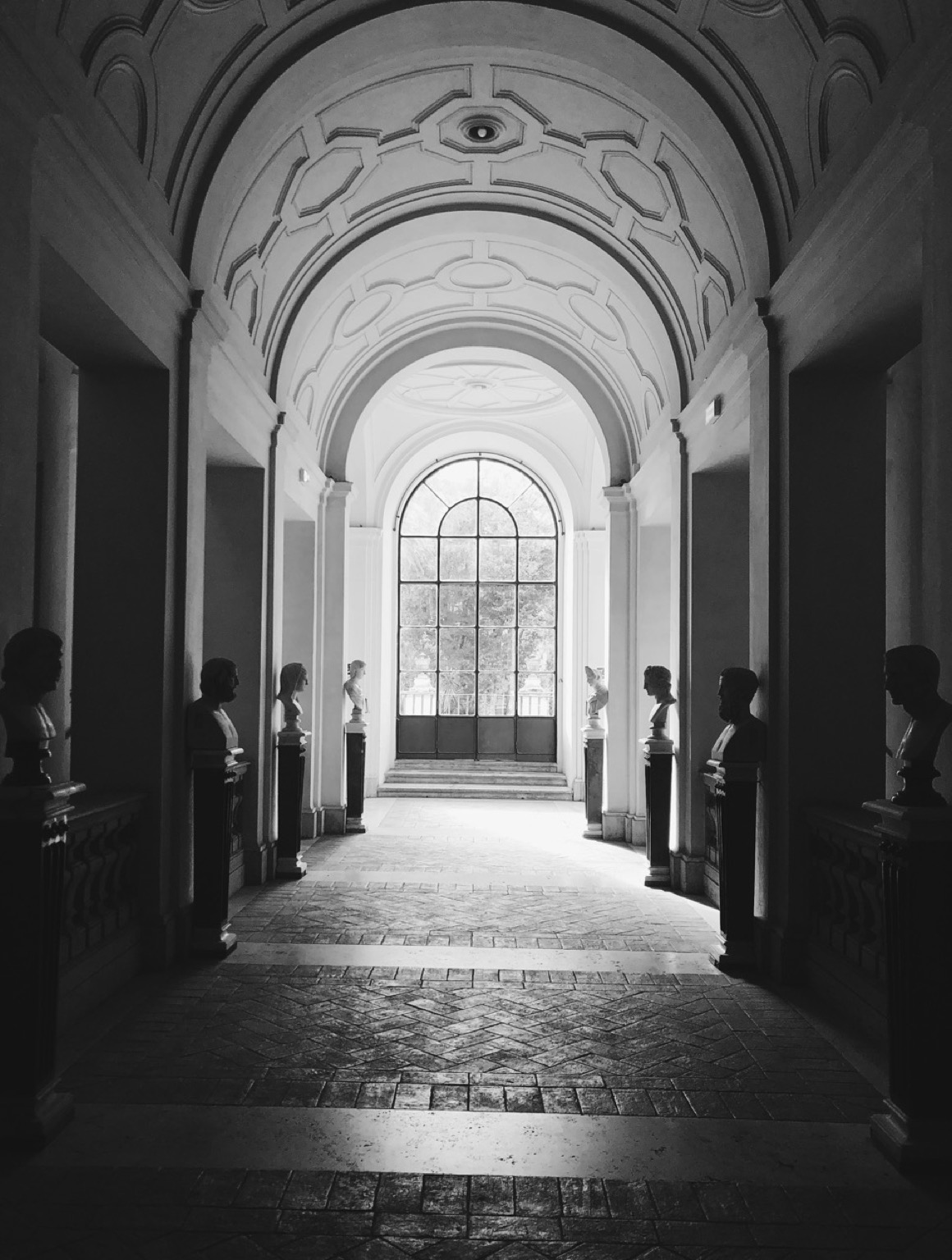 Black and white phography of a corridor in a palace in Rome, Italy. The light comes in through an arc window at the end of the corridor.