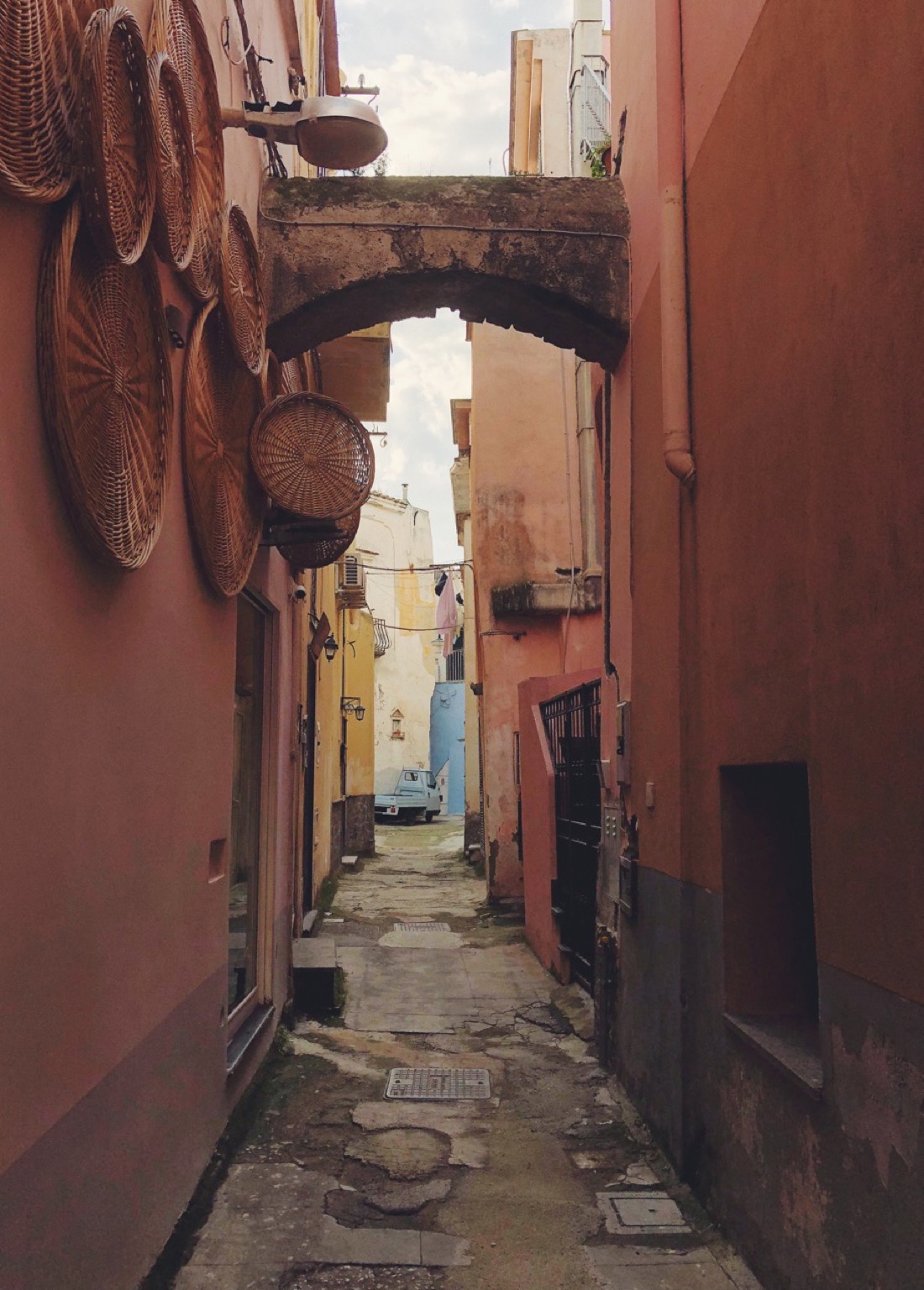 A narrow alley in Naples. Walls are orange and yellow and a small blue car is parked on the end of it.