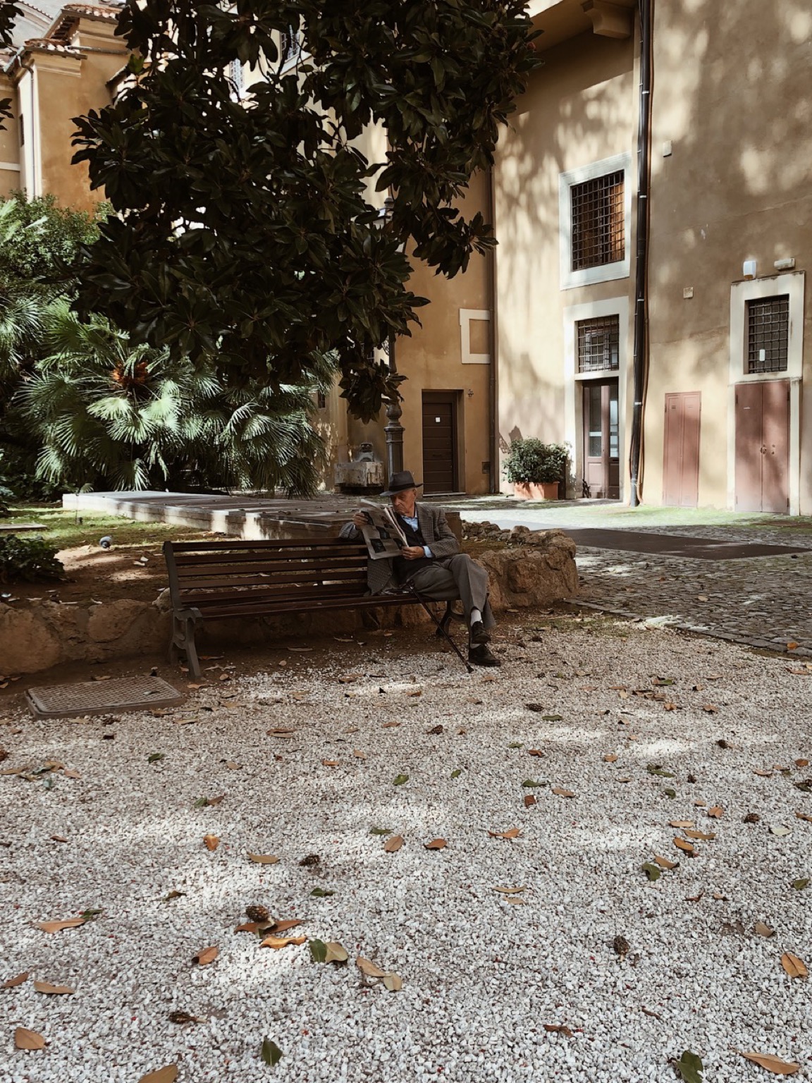 A man is seating on a bench in a park in Rome. He is wearing a costume and a hat while reading a newspaper.